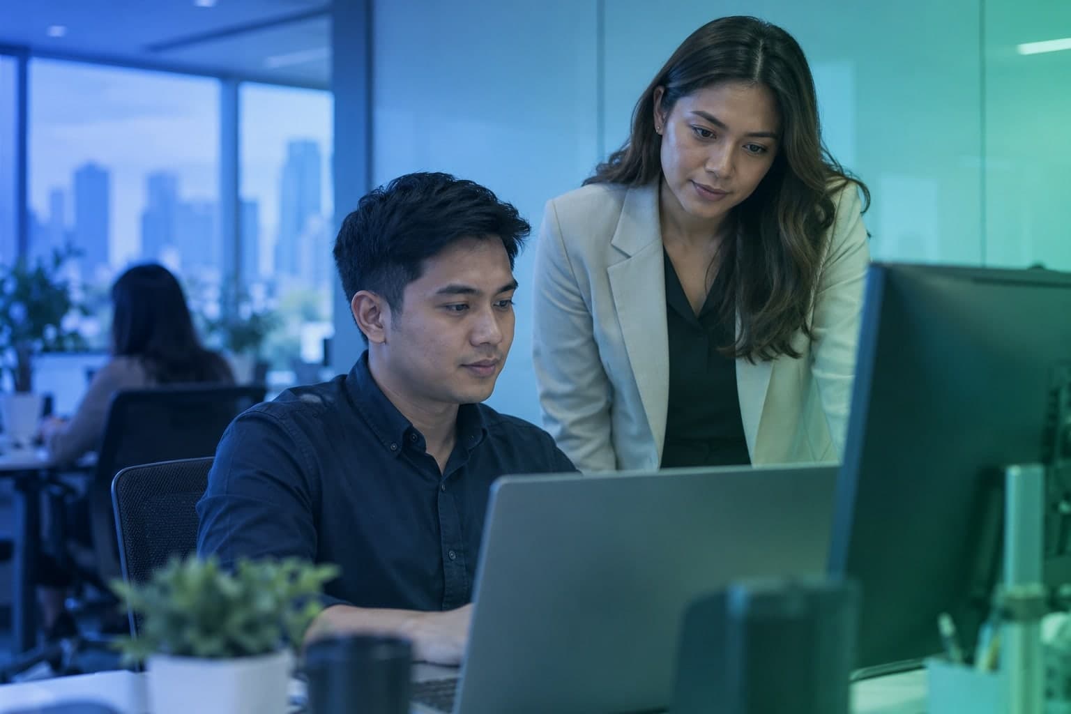 Two Filipino professionals reviewing a laptop and desktop monitor in a blue-and-teal office.